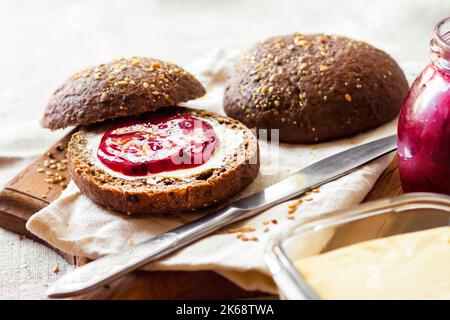 Hausgemachte Roggenbrötchen mit Leinsamen, Sesam und weißen Mohn serviert mit Butter und Jostaberry-Marmelade auf Holzbrett Stockfoto