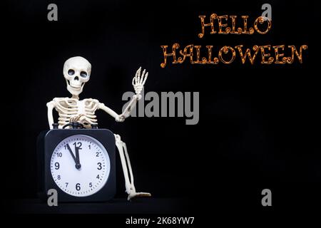 Ein menschliches Skelett winkt mit der Hand und begrüßt einen schwarzen Wecker auf schwarzem Hintergrund mit dem Text Hallo Halloween, Kopierraum. Es ist Hallowee Stockfoto