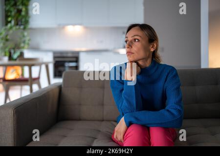 Nachdenkliche Frau sitzt auf einem bequemen Sofa mit erschöpftem und depressiven Ausdruck in der Wohnung Stockfoto