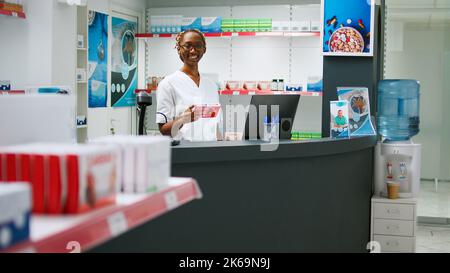 afroamerikanischer Spezialist, der in der Apotheke mit pharmazeutischen Produkten in Regalen arbeitet und Medikamente und Nahrungsergänzungsmittel verkauft. Mitarbeiter, der am Schreibtisch der Drogerie sitzt, um Kunden und Kunden zu helfen. Stockfoto