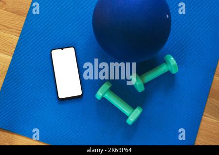 Draufsicht auf modernes Smartphone mit leerem Bildschirm auf Sportmatte mit Ball und Hanteln. Flach legen, mockup. Online-Schulungskonzept. Stockfoto