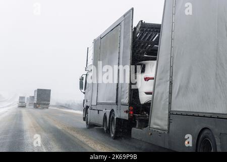 Abschleppwagen Autotransporter Sattelanhänger auf der Autobahn trägt Charge von neuen eingewickelten Elektro-SUVs auf der Autobahn Straße bei starkem Schneefall im Winter. Unternehmen Stockfoto