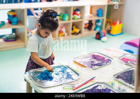 Vorschulkinder stehen am Tisch und legen ihre Hände in das Aquarell, Kindergarten Medium Shot. Hochwertige Fotos Stockfoto