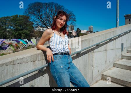 Eine schöne rothaarige junge Frau, die sich an einem sonnigen Tag bequem an einer niedrigen Wand im Freien lehnt. Amerikanisches Flugzeug, Copyspace. Stockfoto