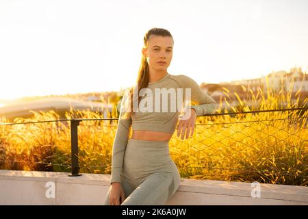 Sportlerin, die nach dem morgendlichen Training in der Sommerstadt sitzt und selbstbewusst auf die Kamera schaut. Aktiver Lebensstil. Fit kaukasischen Weibchen in Sportkleidung bereit zum Joggen. Fitness-Frau bei Sonnenuntergang Stockfoto