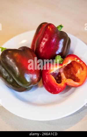Drei selbst angebaute rote Paprika, die sich nur rot färbte, wobei ein Schnitt auf einem weißen Teller in die Hälfte geschnitten wurde. USA. Stockfoto