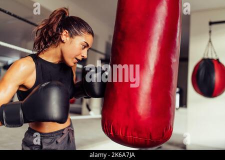 Entschlossene Boxerin, die beim Training im Fitnessstudio einen roten Beutel sticht. Athletische junge Frau, die ihre Schlagtechniken in einer Boxhalle übt. Stockfoto