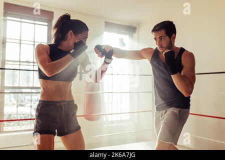 Männlicher Kämpfer, der in einem Boxring eine weibliche Athletin anschlagen kann. Zwei junge Boxer, die in einer Boxhalle trainieren. Stockfoto