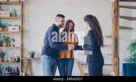 Friendly realtor gibt Schlüssel für junge Paar Käufer von neuen Haus, glückliche Ehegatten umarmen, lächelnden Mann schüttelt die Hände mit Broker macht Deal. Stockfoto