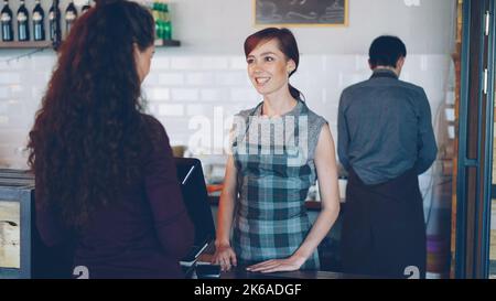 Die Mitarbeiterin des Kaffeehauses spricht mit dem Kunden und verkauft Kaffee zum Mitnehmen an der Theke. Modernes Café- und Getränkekonzept. Stockfoto