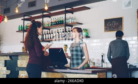 Junge Menschen kaufen in einem netten lokalen Café Kaffee zum Weitertrinken und bezahlen mit dem Smartphone, während freundliche Mitarbeiter Kunden begrüßen, reden und Getränke verkaufen. Stockfoto