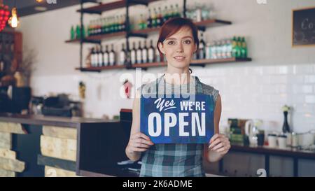 Porträt einer attraktiven selbstbewussten Frau Kleinunternehmen Inhaber halten wir sind offene Zeichen stehen in ihrem Café und lächelnd Blick auf die Kamera. Kaffeehaus im Hintergrund. Stockfoto