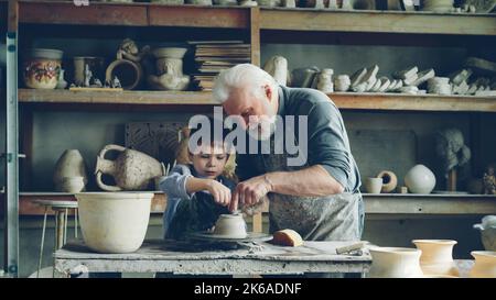 Fürsorglicher Großvater zeigt jungen niedlichen Enkel, wie man mit Lehm am Wurfrad in einer kleinen Werkstatt arbeitet. Töpferei, Familienhobby und Bastelkonzept. Stockfoto