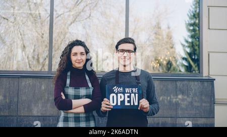 Portrait von attraktiven Freunden Geschäftspartner Eröffnung Restaurant und halten wir sind offen Schild vor dem Fenster vor dem Gebäude. Erfolgreiches Start-up-Konzept. Stockfoto
