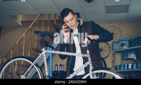 Attraktiver junger Mann fröhlicher Mechaniker ist damit beschäftigt, auf dem Handy zu reden und Fahrradgriff-Bar zusammenzustellen. Menschen-, Wartungs- und Kommunikationskonzept. Stockfoto