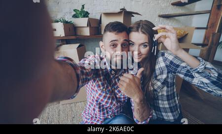 Blickwinkel des glücklichen jungen Paares, das nach dem Kauf einer neuen Wohnung ein Selfie mit den Hausschlüsseln gemacht hat. Junge Menschen halten die Kamera in Posen und küssen sich mit Kisten im Hintergrund. Stockfoto