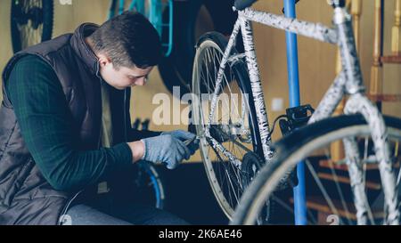 Gutaussehender männlicher Militärangehöriger in Handschuhen repariert das Hinterrad des Fahrrads mit professionellen Werkzeugen. An der Wand hängende Fahrradersatzteile und kleine Werkstatt sind sichtbar. Stockfoto