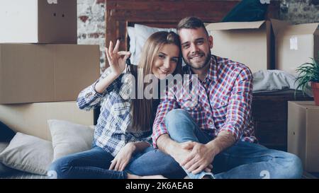Porträt eines glücklichen Ehepaares, das in einem neuen Haus sitzt, Schlüssel hält und mit Kartons herum auf die Kamera schaut. Familienkonzept, Beziehungs- und Umzugskonzept. Stockfoto