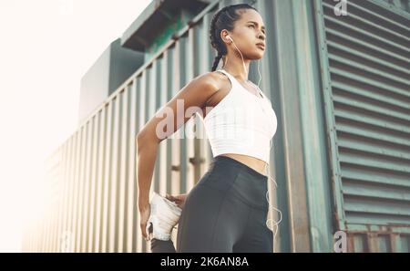 Eine junge hispanische Frau von unten hört Musik mit Kopfhörern und steht auf einem Bein für Quad-Aufwärmstrecken, um Verletzungen zu vermeiden, während Stockfoto