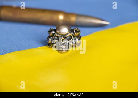 Eine Kugel und ein Ring mit silbernem Schädel liegen auf der blau-gelben Nationalflagge der Ukraine, stoppen den Krieg in der Ukraine, schützen das Land Stockfoto