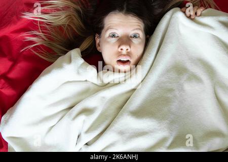Ein junges Mädchen sieht mit Überraschung im Bett auf einem roten Laken aus und ist mit einer weißen Decke bedeckt, schläft und ruht zu Hause im Bett, ein Gefühl der Überraschung und Stockfoto