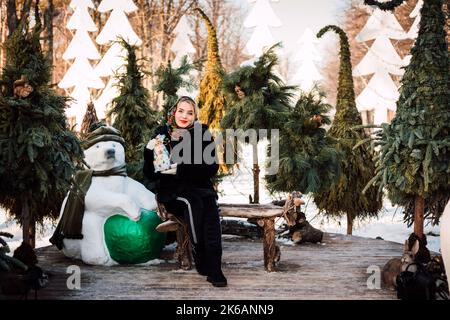 Eine Frau in einem nationalen ukrainischen Kopftuch sitzt auf einer Bank in der Nähe von Weihnachtsbäumen Stockfoto