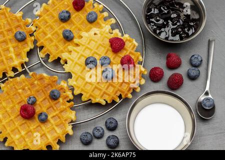 Beeren und Waffeln. Creme und Marmelade in Metallschüsseln. Heidelbeeren im Löffel. Flach liegend. Grauer Hintergrund. Stockfoto