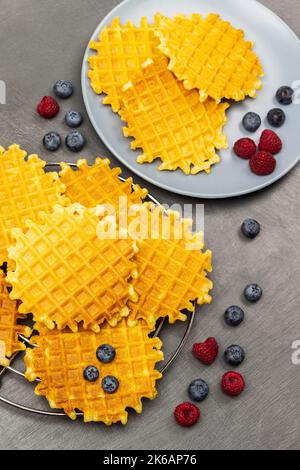 Knusprige Waffeln auf grauem Teller und Metallständer. Himbeeren und Heidelbeeren auf dem Tisch. Flach liegend. Grauer Hintergrund. Stockfoto