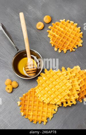 Wiener Waffeln und gelbe Himbeeren auf dem Tisch. Metallschale mit Honig und Holzhonigstäbchen. Flach liegend. Grauer Hintergrund. Stockfoto