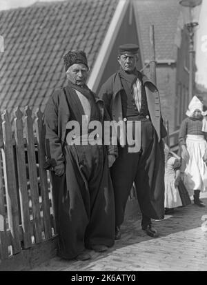 Zwei Männer in traditioneller Kleidung in den Niederlanden c. 1930. Zwei Mädchen stehen hinter ihnen. Männer tragen ein traditionelles Kostüm mit einem langärmeligen Button-up-Hemd zusammen mit einer baggy, plissierten, oft aus Wolle gefertigten Vorderhose, mit silbernen Knöpfen an der quadratischen Frontklappe, Halstuch und Hut – die Form und das Design des Hutes variiert je nach Region. Zu den traditionellen Kostümen gehören in der Regel ein Paar Lederschuhe oder Holzschuhe. Der Stil der traditionellen Kleidung, die in den Niederlanden getragen wurde, begann im 16.. Und 17.. Jahrhundert – ein Vintage-Foto von 1920s/30s. Stockfoto