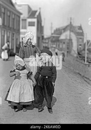 Drei Kinder in traditioneller Kleidung auf einer Straße in den Niederlanden c. 1930. Die traditionellen Kostüme variieren in den 12 niederländischen Provinzen je nach Farbe und Design – Ähnlichkeiten sind das einfache langärmlige Kleid und die Schürze, die von Frauen getragen werden, und die rustikale Arbeitskleidung, die von Männern getragen wird. Die Kleidung für Kinder hat ein ähnliches Design – ein Vintage-Foto von 1920s/30s. Stockfoto