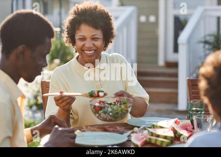 Porträt einer lächelnden afroamerikanischen Frau, die der Familie Essen serviert, während sie gemeinsam im Sonnenlicht im Freien zu Abend gegessen hat Stockfoto