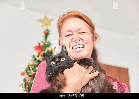 Glückliche, reife hispanische Frau, die breit lächelt, während sie ihre niedliche Katze zu Weihnachten in ihrem Haus hält. Selektiver Fokus auf das Gesicht des Tieres. Stockfoto