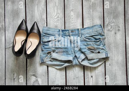 Denim-Shorts und schwarze Schuhe mit hohem Absatz liegen auf einem weißen rustikalen Tisch, trendige Denim-Kleidung Stockfoto