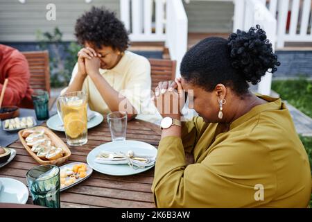 Seitenansicht Porträt einer schwarzen jungen Frau, die während des Familientreffens am Tisch im Freien betet Stockfoto