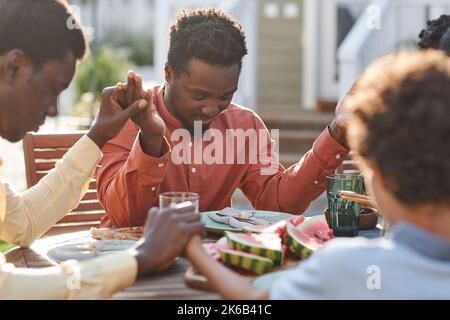 Porträt eines jungen schwarzen Mannes, der im Freien am Tisch Gnade sagt, während die Familie zusammenkommt und die Hände im Sonnenlicht hält Stockfoto