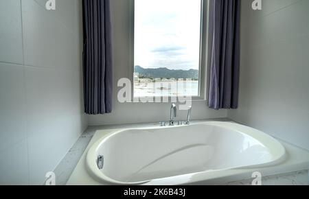 Nahaufnahme der Badewanne mit Blick auf das Meer und die Berge im Hintergrund. Es gibt weiße Wände, Es gibt große offene Fenster mit Blick auf das Meer, Berge und Stockfoto