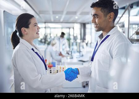 Zwei verschiedene professionelle Wissenschaftler schütteln sich die Hände, während sie blaue Handschuhe und Laborkittel tragen. Zwei Ärzte stimmen zu und gratulieren einem Stockfoto