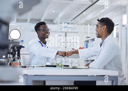 Zwei lächelnde medizinische Wissenschaftler sitzen in einem Labor zusammen und stoßen mit der Faust. Afroamerikanische und gemischtmedizinische Fachkräfte feiern Stockfoto