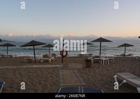 Sandstrand eines Ferienortes mit Plastikteppich, Plastikliegen und Strohsonnenschirmen zur Sonnenaufgangszeit. Stockfoto