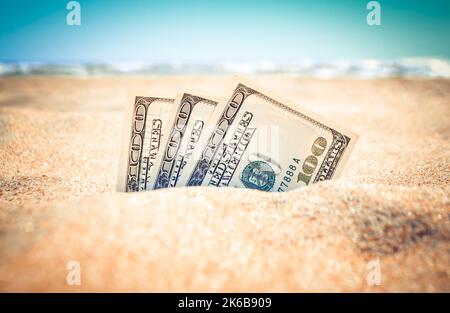 Die mit Sand bedeckten Gelddolare liegen am Sandstrand in der Nähe der Meereswellen am sonnigen Sommertag aus nächster Nähe. Geld wächst aus dem Boden. Konzept Finanzierung Geld Urlaub Relax Urlaub. Stockfoto