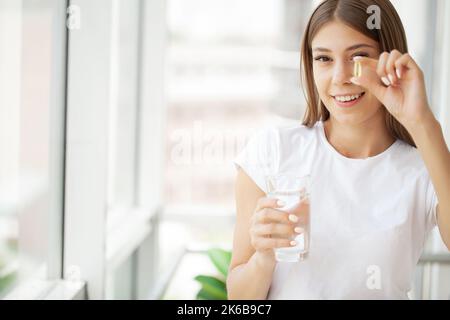 Frau, die eine Pille mit Cod Liver Oil Omega-3 eingenommen hat und morgens Ein Glas frisches Wasser hält Stockfoto