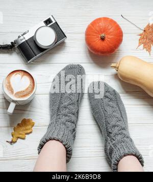 Beine eines Mädchens in gestrickten Socken auf einem Holzhintergrund neben Kürbissen, einer Tasse Kaffee, einer Kamera und Herbstblättern. Gemütliches Herbstkonzept. Stockfoto