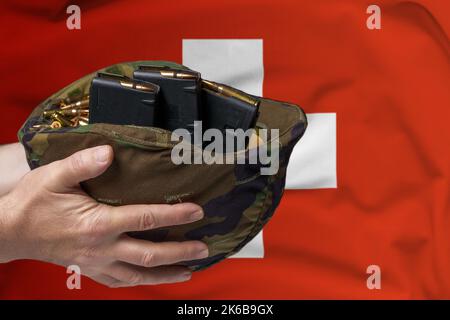 Ein Militärhelm mit Patronen und Zeitschriften für ein Gewehr in den Händen eines Mannes vor dem Hintergrund der Schweizer Flagge. Das Konzept von Stockfoto