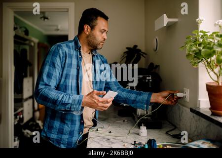 Mann, der das Smartphone-Ladegerät in die Steckdose gesteckt hat, während er zu Hause in der Nähe der Küchentheke stand Stockfoto