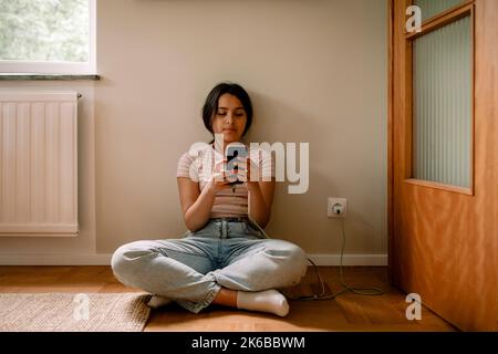 Mädchen mit Smartphone, während sie zu Hause mit gekreuzten Beinen auf dem Boden gegen die Wand sitzt Stockfoto
