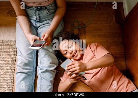 Lächelnder Junge, der zu Hause Smartphone mit seiner Schwester benutzt, während er sich auf dem Boden niederlegt Stockfoto