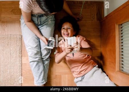 Direkt über der Aufnahme von glücklichen Geschwistern, die zu Hause Smartphones auf Hartholzboden benutzen Stockfoto