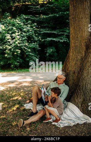 Großvater liest dem Enkel das Märchbuch vor, während er unter dem Baum im Park sitzt Stockfoto