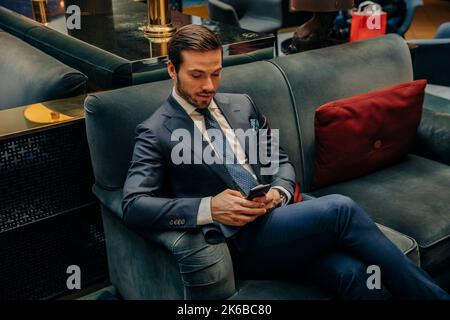 Der junge Geschäftsmann hat SMS über das Smartphone geschickt, während er auf dem Sofa in der Hotellounge sitzt Stockfoto